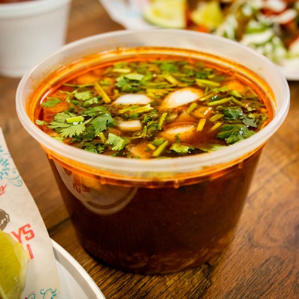 Rico's Tacos' birria consommé in a clear plastic cup, garnished with cilantro and onions.